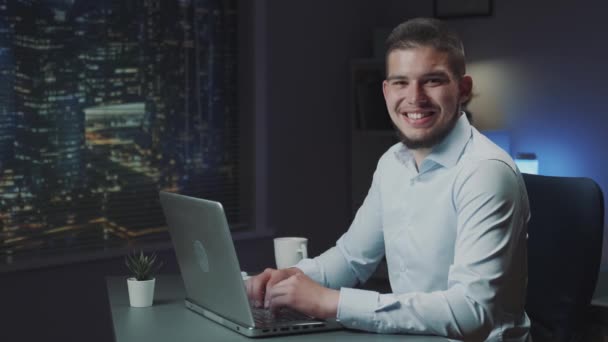 Hombre guapo con barba sonriendo a la cámara mientras trabaja en el ordenador — Vídeos de Stock