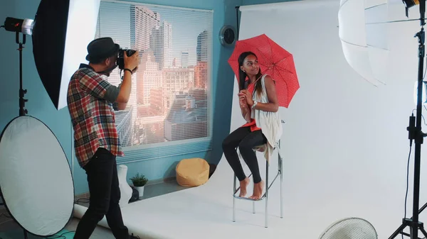 スタイリッシュなアフリカのモデルは、ファッション雑誌の写真撮影のためのバーの高い椅子の上に赤い傘でポーズ — ストック写真