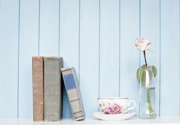 Old books, china cup and rose in the bottle on bookshelf — Stock Photo, Image