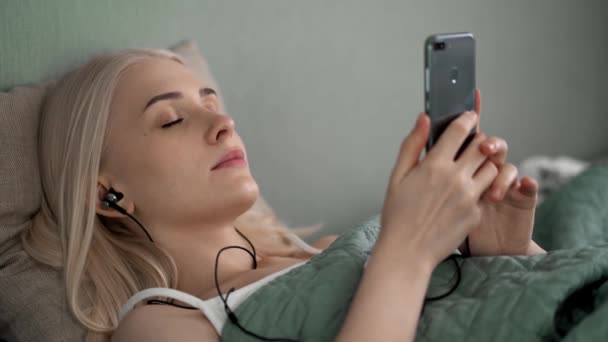Gyönyörű lány fekszik az ágyon, zenét hallgat fejhallgatóval. — Stock videók