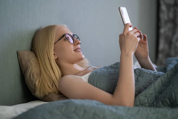 Smiling woman looking at mobile phone lying down on white bed. Happy blond young woman using cellphone at home.