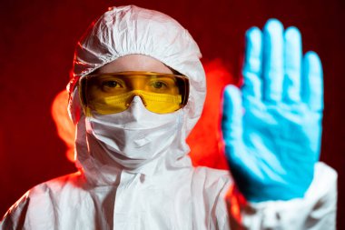 Koruyucu kıyafetli bir doktor kırmızı arka planda dur işareti gösteriyor. Coronavirüs salgını konsepti COVID-19, virüsün bulaşma tehlikesi