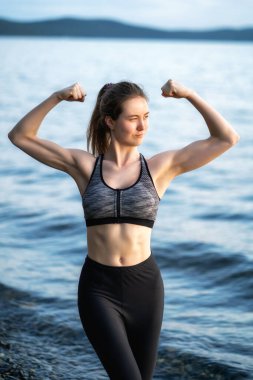 Genç ve güzel kadın vücut geliştirici poz veriyor ve gölde kaslarını gösteriyor.