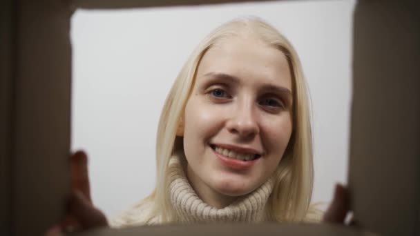 Vista desde el interior de la caja de cartón en la joven mujer blanca alegre que mira en ella y sonriendo. — Vídeos de Stock