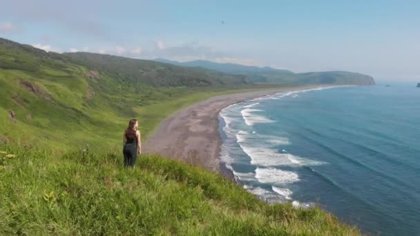 Jovem mulher olhando para o oceano, de pé em um penhasco alto acima da praia de areia. — Vídeo de Stock