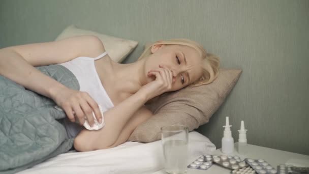Uma jovem menina bonita está doente, deita-se no auto-isolamento na cama, limpa o nariz — Vídeo de Stock