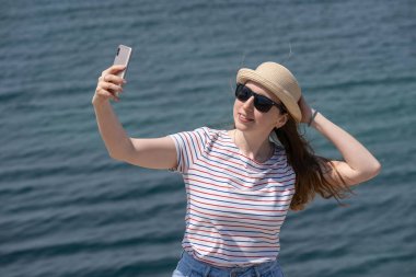 Bir kadın şapkasını eliyle tutuyor ve telefonunu gölde kameraya çekiyor. Rüzgarlı bir hava. Açık hava eğlencesi, büyük şehirden uzakta..