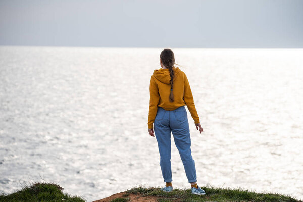 A woman traveler in a bright yellow sweatshirt and loose jeans stands on the seashore and looks into the distance. Freedom and a sense of tranquility.