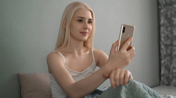 Una foto auténtica de una joven rubia está haciendo una videollamada o selfie con un teléfono inteligente antes de dormir en un acogedor Imagen de stock