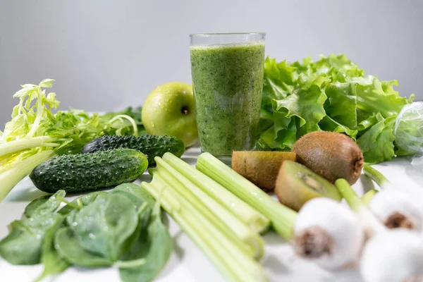 Diyet detoks smoothie beyaz bir masada duruyor. Çevrelerinde (cennette) yeşil sebzeler, meyveler vardır. Vitaminler ve sağlıklı yiyecekler.
