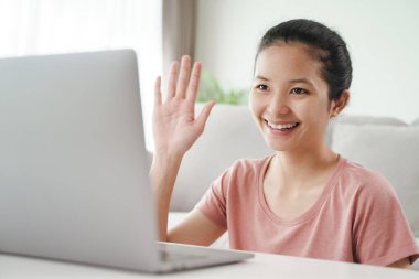 Genç Asyalı kadın online video konferansı için dizüstü bilgisayar kullanıyor. El sallayarak oturma odasında selam veriyor..