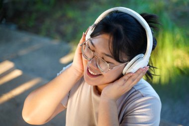 Neşeli, genç Asyalı bir kadın kulaklık takıyor, gözlerini kapıyor ve neşeli bir ifadeyle müzikten zevk alıyor. Rahatlama, zihinsel sağlık ve modern ses teknolojisi yaşam tarzı kavramı.