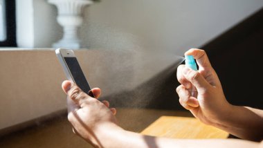 Genç Asyalı adamın eli akıllı telefonlardaki mikropları öldürmek için alkol spreyi püskürtüyor mikroplara karşı ilk koruma, salgın sırasında elektronik aletlerin temizlenmesi ya da dezenfektanların temizlenmesi..