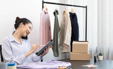 Genç Asyalı bir kadın akıllı telefon kullanarak müşteriyle sohbet ediyor ve siparişi onaylamak için tablet kullanıyor, çevrimiçi kıyafet satış işi, kıyafet satışı veya internet üzerinden satış işi.