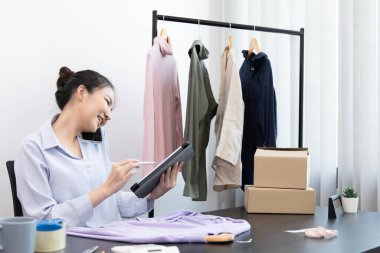 Genç Asyalı bir kadın akıllı telefon kullanarak müşteriyle sohbet ediyor ve siparişi onaylamak için tablet kullanıyor, çevrimiçi kıyafet satış işi, kıyafet satışı veya internet üzerinden satış işi.