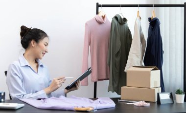 Genç Asyalı kadınlar müşteri ihtiyaçlarını karşılamak için tabletle kıyafet stoklarını kontrol ediyorlar, satmadan önce ürünleri sayıyorlar, çevrimiçi giyim satış işleri, giysi satma veya İnternet üzerinden satış işleri.