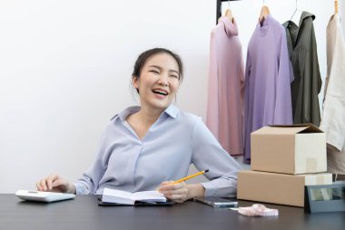 Yüzü gülen Asyalı kadın müşterilerin moda siparişlerini not alıyor, iyi hissettiriyor, sosyal medyada kıyafet satma işi ya da internetten satış, sosyal mesafede yeni iş..