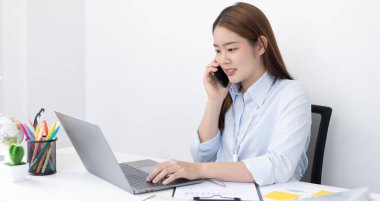 Asyalı kadın çalışan telefonda konuşuyor ve gülümseyen bir yüzle dizüstü bilgisayarda çalışıyor, ofis çalışanları iş ve telefon konuşmaları, telefonda iş konuşmaları..