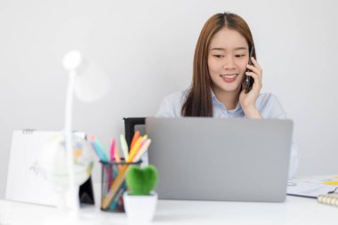 Asyalı kadın çalışan telefonda konuşuyor ve gülümseyen bir yüzle dizüstü bilgisayarda çalışıyor, ofis çalışanları iş ve telefon konuşmaları, telefonda iş konuşmaları..