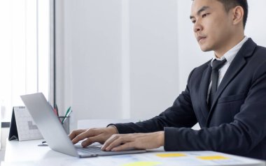Genç Asyalı ofis çalışanı finansal verileri analiz etmek ve çalışmalarını özetlemek için dizüstü bilgisayara basıyor. Ofis çalışanları, siyah takım elbiseli yakışıklı erkek çalışanlar..