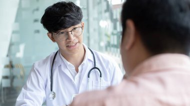 Beyaz cüppeli genç Asyalı bir adam bir adamla konuşuyor, doktorlar hastalıklara teşhis koyuyor ve hastane muayene odasındaki hastalara tavsiye ya da tedavi çözümleri sunuyor..