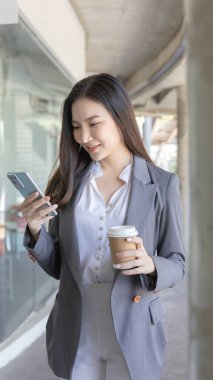 Gri takım elbiseli genç Asyalı kadın elinde bir telefon ve kağıt kahve kupası tutuyor, iş kadınları iş ortaklarıyla sohbet ederek veya konuşarak iletişim kurmak için akıllı telefon kullanıyor, İnternet medyası, VDO çağrısı.