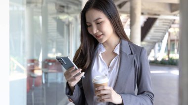 Gri takım elbiseli genç Asyalı kadın elinde bir telefon ve kağıt kahve kupası tutuyor, iş kadınları iş ortaklarıyla sohbet ederek veya konuşarak iletişim kurmak için akıllı telefon kullanıyor, İnternet medyası, VDO çağrısı.