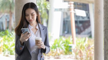 Gri takım elbiseli genç Asyalı kadın elinde bir telefon ve kağıt kahve kupası tutuyor, iş kadınları iş ortaklarıyla sohbet ederek veya konuşarak iletişim kurmak için akıllı telefon kullanıyor, İnternet medyası, VDO çağrısı.