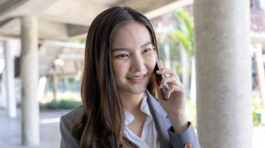 Gri takım elbiseli genç Asyalı kadın bir ofis binasının altında duruyor telefonla konuşuyor, cep telefonu kullanıyor, iş görüşmeleri yapıyor gri takım elbiseli, gülümseyen bir kadın..