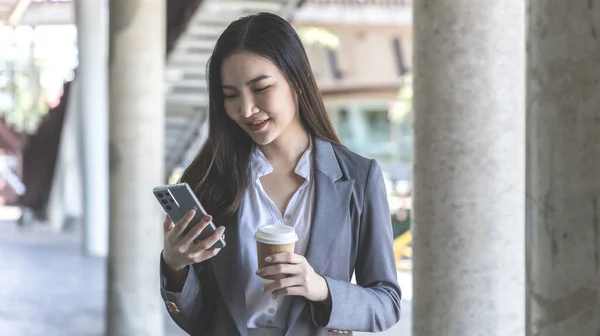 Young Asian woman wearing a gray suit is holding a phone and a paper coffee mug, Business women use smartphones to communicate by chatting or talking with business partners, Online media, VDO call.