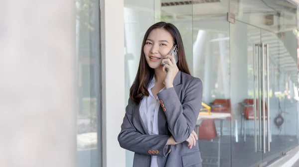 Young Asian female employee in a gray suit stands under the building of an office building talking on the phone, Talking using a mobile phone, Business communication, Smiling woman in a gray suit.