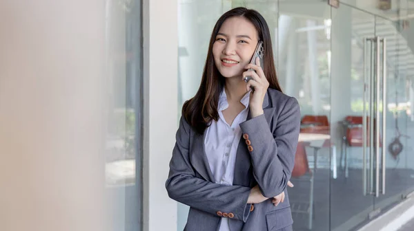 Young Asian female employee in a gray suit stands under the building of an office building talking on the phone, Talking using a mobile phone, Business communication, Smiling woman in a gray suit.