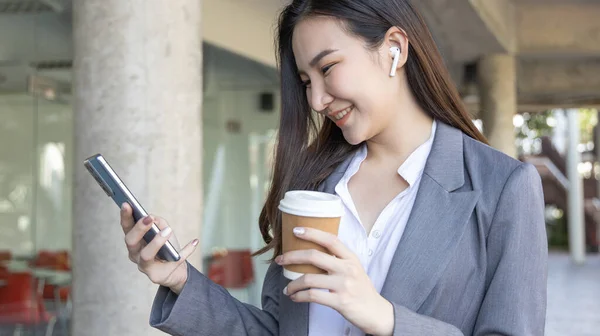 Young Asian woman wearing a gray suit is holding a phone and a paper coffee mug, Business women use smartphones to communicate by chatting or talking with business partners, Online media, VDO call.