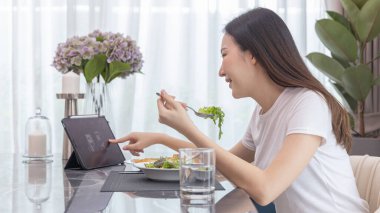 Taze salata yiyen ve evde yemek masasında tablet kullanan Asyalı kadın, vitamin ve mineral, düşük kalorili ve yüksek lifli diyetler, sağlıklı yiyecekler, aperatifler açısından zengin sebze salataları var..