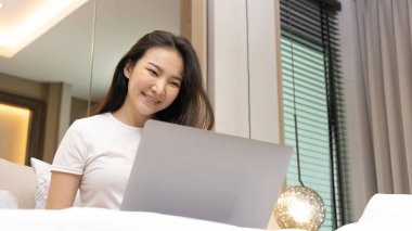 Asyalı bir kadın, sabah kalktıktan sonra evdeki modern yatak odasında dizüstü bilgisayar kullanıyor. Laptop teknolojisi, uyandırma aktiviteleri ve mutlu bir yaşam tarzı kullanıyor..
