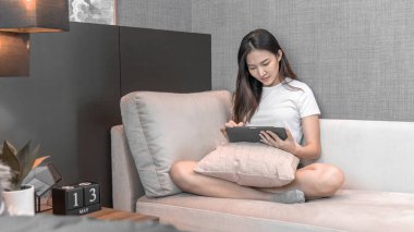 Asyalı bir kadın hafta sonları evinde kanepede oturup, teknolojiyi ve interneti iletişim kurmak, eğlenmek ve rahatlamak, internete girmek, evinde rahat bir yaşam sürmek, yaşam tarzı için kullanıyor..