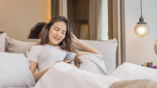 Asian woman uses mobile phone in beautiful modern bedroom at home after she wakes up in the morning, Entertaining or relaxing using mobile phone technology, Wake-up activities, Happy lifestyle.