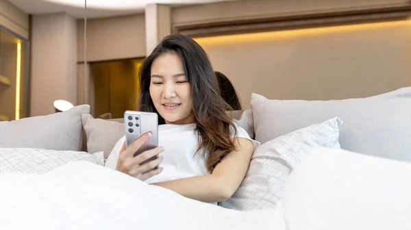 Asian woman uses mobile phone in beautiful modern bedroom at home after she wakes up in the morning, Entertaining or relaxing using mobile phone technology, Wake-up activities, Happy lifestyle.
