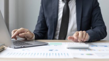 İşadamı kazancını hesaplamak için bir hesap makinesine basar ve bilgisayarını özel ofisi Vergi ve Muhasebe Bilgileri 'nde şirketinin mali istatistiklerini çizmek için kullanır..