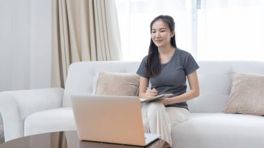Not defterine not alan Asyalı kadın evde dizüstü bilgisayar, görüntülü sohbet, online iletişim, Evde kal, Yeni Normal, Covid-19 Coronavirus, Sosyal Mesafe, Çevrimiçi Öğren.