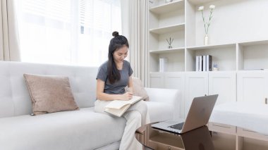 Not defterine not alan Asyalı kadın evde dizüstü bilgisayar, görüntülü sohbet, online iletişim, Evde kal, Yeni Normal, Covid-19 Coronavirus, Sosyal Mesafe, Çevrimiçi Öğren.