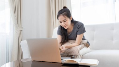 Kanepede oturan Asyalı kadın, ödeme veya çevrimiçi işlemler, mali işlemler ve internet güvenliği için kredi kartı ile online alışveriş yapmak için dizüstü bilgisayar kullanıyor..