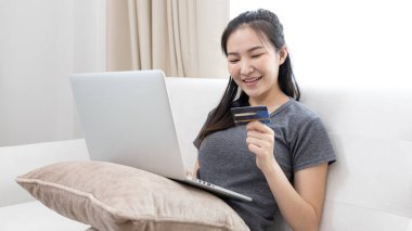 Kanepede oturan Asyalı kadın, ödeme veya çevrimiçi işlemler, mali işlemler ve internet güvenliği için kredi kartı ile online alışveriş yapmak için dizüstü bilgisayar kullanıyor..