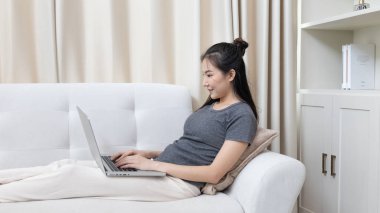 Kanepede oturan Asyalı kadın, ödeme veya çevrimiçi işlemler, mali işlemler ve internet güvenliği için kredi kartı ile online alışveriş yapmak için dizüstü bilgisayar kullanıyor..