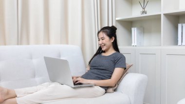 Kanepede oturan Asyalı kadın, ödeme veya çevrimiçi işlemler, mali işlemler ve internet güvenliği için kredi kartı ile online alışveriş yapmak için dizüstü bilgisayar kullanıyor..