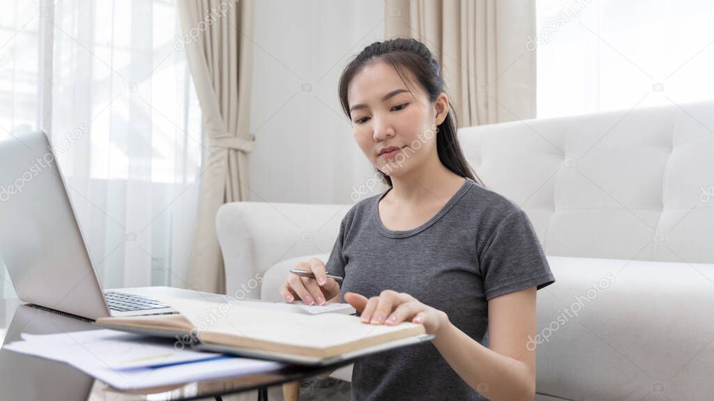 Asian woman working at home on weekends, Press the calculator to calculate the income - expenses of the house and use the laptop to record the information and check the accuracy, Home lifestyle.