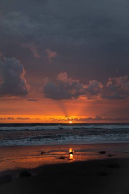 Bali 'de okyanusun kenarında inanılmaz güzel bir gün batımı. Parlak güneş suda batıyor. Güneş ışınları bulutların arkasından süzülüyor ve gün batımının suda güzel bir yansıması.