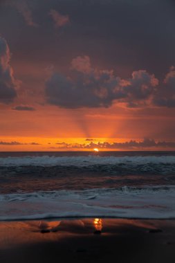 Bali 'de okyanusun kenarında inanılmaz güzel bir gün batımı. Parlak güneş suda batıyor. Güneş ışınları bulutların arkasından süzülüyor ve gün batımının suda güzel bir yansıması.