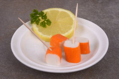 Surimi tahta kazmalarla bir tabak içinde limon dilimi ve gri bir arka planda maydanozla yakın plan çekilir. 