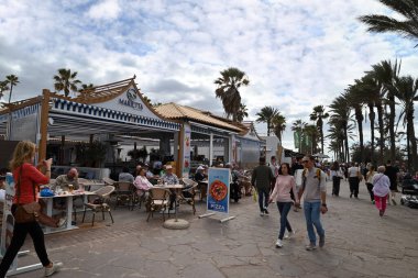 Müşteriler bir bar-restoran terasında oturuyor ve turistler Tenerife adasındaki Playa de Las Americas 'ta geziniyorlar.